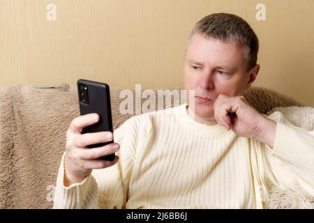 Un europeo in un maglione bianco siede su un divano con un telefono in mano. Un uomo guarda pensieroso in uno smartphone, poggiando la testa sulla mano. Foto Stock
