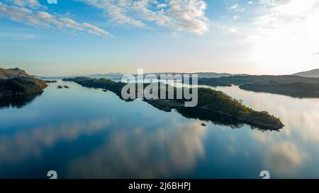 L'acqua è come uno specchio, il cielo blu con piccole nuvole si riflette in esso, il sole tramonta lateralmente, nel mezzo di una lunga isola. Foto Stock