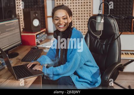 Ingegnere del suono di charme con un bel sorriso toothy guardando la fotocamera mentre si lavora su un computer portatile in uno studio di registrazione musicale Foto Stock