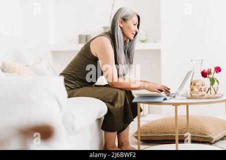 Donna asiatica matura che lavora con il laptop mentre si siede sul divano a casa Foto Stock