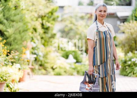 Donna asiatica matura che indossa grembiule che lavora nel suo giardino il giorno d'estate Foto Stock