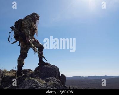 Il concetto di guerra moderna - un mercenario cecchino ispeziona l'area da una montagna alta. Un soldato in mimetizzazione che si fonde con l'ambiente Foto Stock