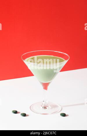 frullato verde in bicchiere da cocktail martini con pillole di alghe spirulina su sfondo rosso. cura di sé e minimo sano concetto di vita partito. spazio copia Foto Stock