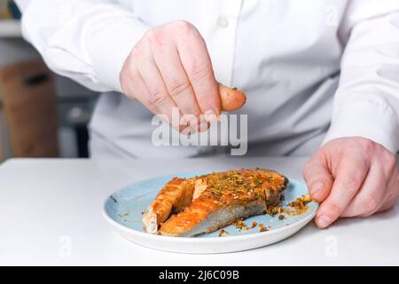 Il cuoco cosparge la bistecca di pesce rosso con spezie. La bistecca di salmone si trova su un piatto Foto Stock