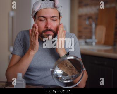 L'idea di bellezza degli uomini. Giovane uomo bello in cucina in concetto di igiene. Archetto, specchio sul tavolo, detergente per imperfezioni cutanee Foto Stock