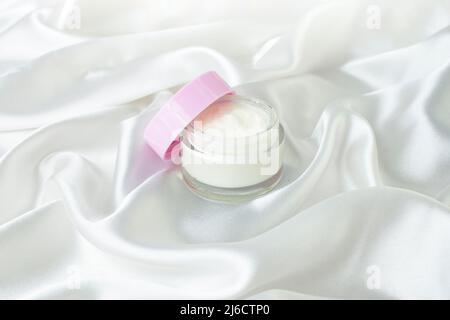 Vaso in vetro crema con coperchio rosa aperto, su sfondo bianco satinato. Cura della pelle semplice mock up con onde seriche podio per la visualizzazione Foto Stock