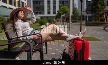 Bella donna caucasica in un cappello e pantaloncini si siede su una panca che allunga le gambe su una valigia rossa e in attesa di un taxi. Ragazza in attesa di partenza Foto Stock