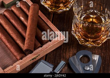 Scatola con sigari cubani, accendisigari e taglierina su un vecchio tavolo in legno. Due bicchieri di whiskey o alcol sullo sfondo. Foto Stock