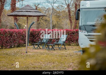 Due sedie a sdraio vuote di fronte ad una moderna e elegante casa di moto parcheggiata in un bel campeggio circondato da lussureggianti piante rosse. Campeggio moderno con camma Foto Stock