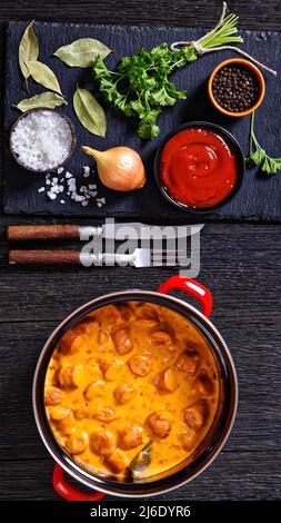 nakkikastike, salsicce finlandesi, amalgia in salsa di pomodoro cremosa in pentola rossa su tavola di legno scuro con ingredienti, vista verticale dall'alto, posa piatta Foto Stock