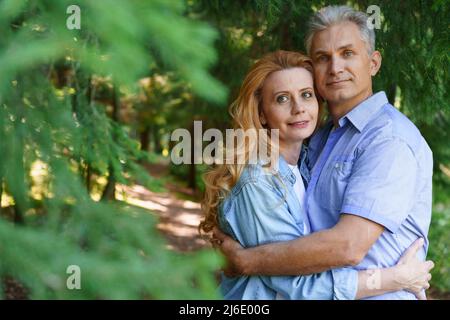 Vecchiaia, relazioni e persone concetto di coppia anziana felice abbraccia nel parco della città sullo sfondo di alberi in abiti casual chiaro. La gente felice passa il tempo insieme in una giornata di sole in estate Foto Stock