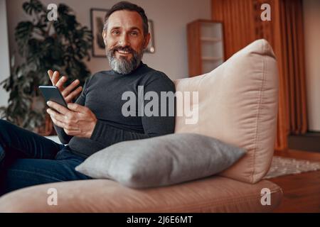 Uomo sorridente con telefono cellulare seduto in una comoda poltrona Foto Stock