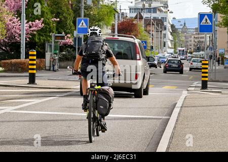 Basilea, Svizzera - Aprile 2022: Persona in bicicletta a casa dal lavoro dietro un'auto in una strada alla periferia del centro della città Foto Stock