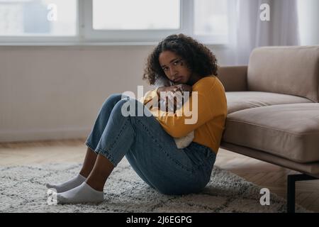 Giovane donna nera con depressione seduta sul pavimento e cuscino abbracciato a casa Foto Stock
