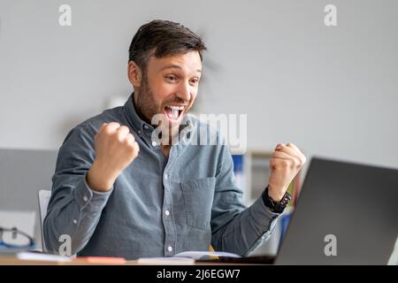 Fortuna di affari. Un uomo d'affari maturo e pieno di gioia che utilizza un notebook e batte i pugni, festeggia grandi notizie, seduto in ufficio Foto Stock