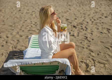 Signora bionda che si gode mohito cocktail e guardare via sulla spiaggia vuota Foto Stock
