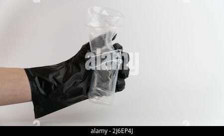 Mano di primo piano di una donna indossare guanti di plastica nera schiacciando una tazza di plastica su sfondo bianco in studio, concetto di riciclaggio. Foto Stock