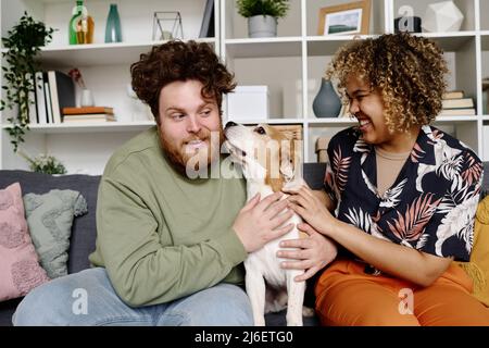 Coppia multietnica divertirsi sul divano con il loro cane, giocando con l'animale domestico e ridendo Foto Stock