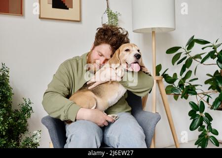 Giovane uomo portico seduto su poltrona in camera con piante che abbracciano il suo cane e rilassano Foto Stock