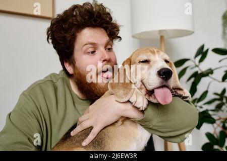 Giovane uomo bearded che tiene il cane nelle sue braccia ed attacca la sua linguetta appena come il suo cane Foto Stock