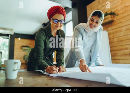 Felici architetti musulmani che lavorano su disegni blueprint in un ufficio moderno. Due imprenditrici creative che progettano un nuovo progetto innovativo. Designazione femminile Foto Stock