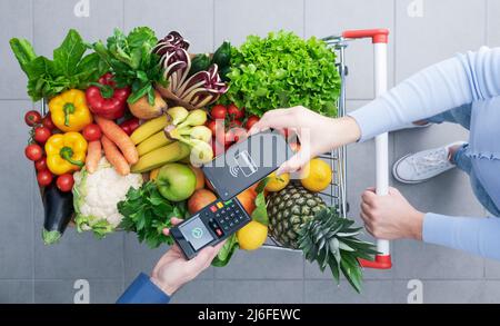 Donna che spinge un carrello completo per la spesa e paga per i generi alimentari con il suo smartphone, concetto di pagamenti mobili, vista dall'alto Foto Stock