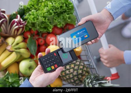 Donna che spinge un carrello completo per la spesa e paga per i generi alimentari con il suo smartphone, concetto di pagamenti mobili, vista dall'alto Foto Stock