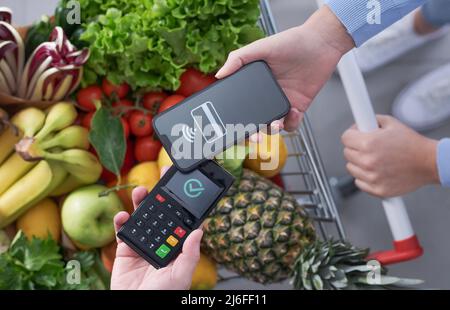 Donna che spinge un carrello completo per la spesa e paga per i generi alimentari con il suo smartphone, concetto di pagamenti mobili, vista dall'alto Foto Stock