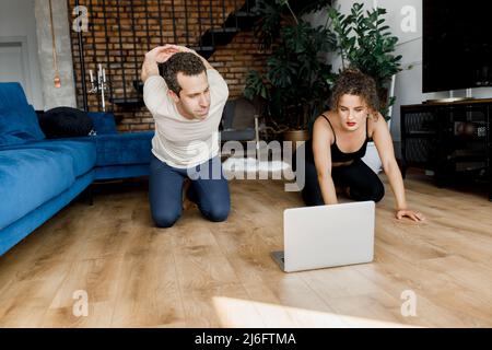 Calzare giovane uomo facendo esercizio sportivo a casa con il computer portatile sdraiato sul pavimento con la ragazza che sostiene il ragazzo di aiuto, donna di allenamento sportivo marito a. Foto Stock