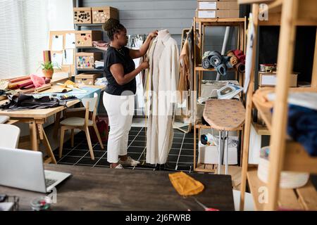 Giovane afroamericana su misura davanti al manichino con cappotto e misure con nastro di misurazione in officina Foto Stock