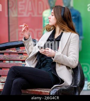 Una bella giovane donna seduta all'aperto rilassarsi al caldo sole primaverile mentre fuma una sigaretta nel centro di Dundee, in Scozia Foto Stock