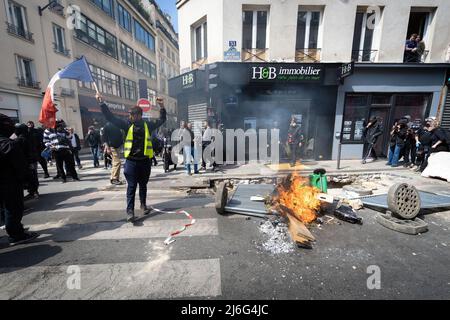 Un manifestante giallo con una bandiera francese cammina accanto a mobili di strada brucianti che è stato messo a fuoco per impedire alla polizia di andare avanti. Migliaia di persone si sono svolte per i raduni del giorno di maggio. I manifestanti hanno marciato attraverso la città chiedendo il progresso sociale in una società ansiosa per il futuro del pianeta e di un mondo in pace. Storicamente il 1st maggio segna la Giornata Internazionale del lavoro che commemora i lavoratori e la classe operaia. Foto Stock