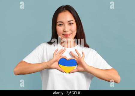 Bella giovane donna asiatica con cuore di carta nei colori della bandiera Ucraina su sfondo blu Foto Stock