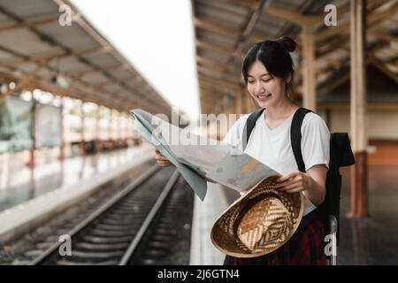 Giovane donna viaggiatore con zaino in cerca di mappa in attesa di treno, saccopelatore asiatico sulla piattaforma ferroviaria alla stazione ferroviaria. Vacanza, viaggio Foto Stock