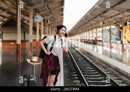 Bella giovane donna asiatica con uno zaino usa il telefono mentre si trova vicino al treno ferroviario sulla piattaforma. Concetto estate di viaggio economico Foto Stock