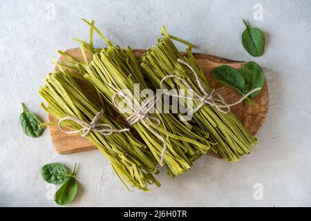 Fasci di fettuccine di pasta cruda con spinaci su tagliere in legno e fondo in calcestruzzo Foto Stock