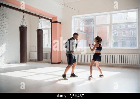 Un giovane pugile attraente insegna le sue tecniche di pugilato fidanzata in un loft attrezzato per l'allenamento di boxe. Foto Stock