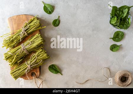 Fasci di fettuccine di pasta cruda con spinaci su tagliere in legno e fondo in calcestruzzo Foto Stock