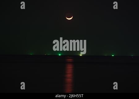 La lunga esposizione della Luna sopra un mare, con il sentiero al chiaro di luna sopra l'acqua è chiaramente visibile insieme alle luci di navi da pesca lontane Foto Stock