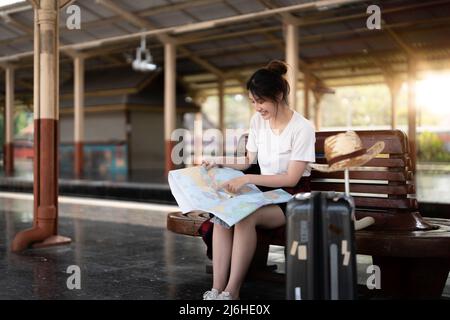 Felice giovane asiatica viaggiatore o zaino in spalla utilizzando mappa scegliere dove viaggiare con i bagagli alla stazione ferroviaria, vacanza estiva concetto di viaggio Foto Stock