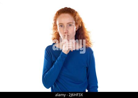 Ritratto interno di giovane donna rossa con capelli ricci lunghi guarda direttamente nella macchina fotografica, sorridendo e mostra il gesto del silenzio, isolato su dorso arancione Foto Stock