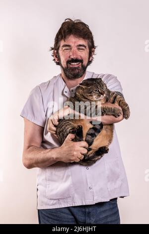 Veterinario con un gatto in braccio, con una giacca bianca. Gatto con strisce. Uomo di mezza età con barba. Sfondo bianco. Concetto cura animali. Foto Stock