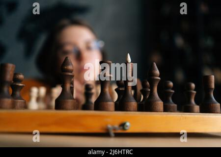 Fuoco selettivo di giovane donna che indossa occhiali eleganti pensando a scacchi muoversi mentre si siede su pavimento di legno in camera oscura. Foto Stock