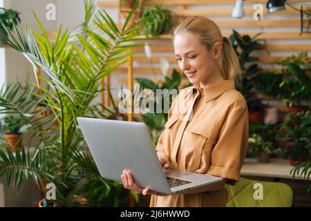 Ritratto di grazioso giovane imprenditore floreale proprietario di negozio in piedi con notebook tenendo in mano tra piante verdi, guardando sullo schermo. Foto Stock