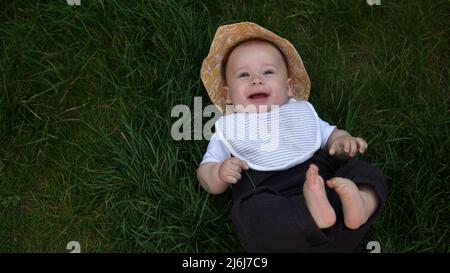 Piccolo bambino neonato felice in estate panama cappello cadere giù posa su erba a piedi nudi in estate Sunny Day. Bambino bambino bambino ragazzo Smacinatura faccia guardare Foto Stock