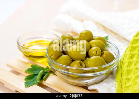 Molte olive verdi intere e olio d'oliva nelle ciotole di vetro su sfondo di legno chiaro in cucina. Foto Stock