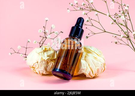 Bottiglia di vetro marrone con olio essenziale e contagocce per la cura della pelle e massaggio su sfondo rosa pastello con fiori. Foto Stock