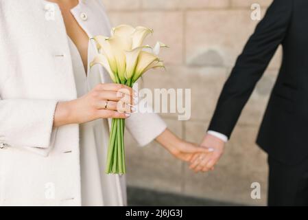 La sposa tiene la mano dello sposo. Bouquet di gigli di calla bianca nelle mani di una donna in un cappotto bianco Foto Stock