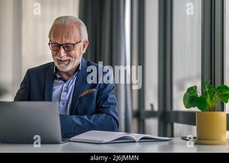 Uomo d'affari anziano sorridente che lavora su un laptop. Indossando formals professionali maschili senior mentre si utilizza un dispositivo portatile. È in tuta seduto alla scrivania in Foto Stock