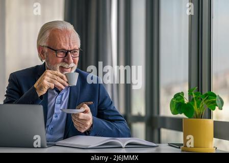 Sorridente professionista anziano che ha un caffè. Uomo d'affari senior che lavora su un notebook alla scrivania. Indossa formali e occhiali in ufficio. Foto Stock